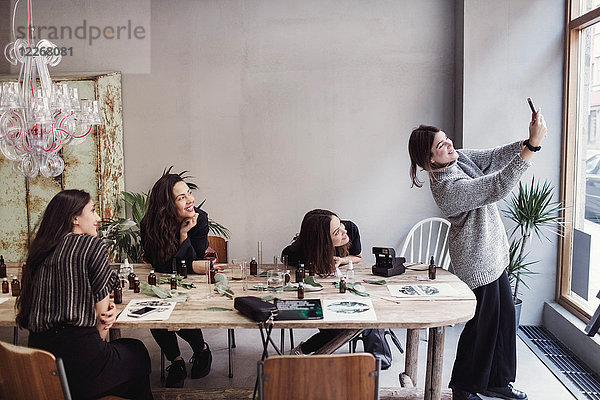 Lächelnde Besitzerin nimmt Selfie mit weiblichen Mitarbeitern am Tisch in der Parfümwerkstatt auf