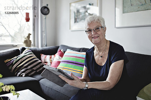 Porträt einer selbstbewussten Seniorin mit digitalem Tablett auf Sofa im heimischen Wohnzimmer