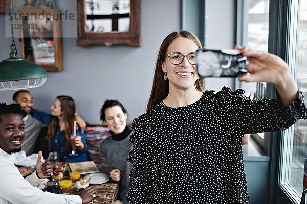 Lächelnde junge Frau nimmt Selfie mit Freunden im Restaurant mit.