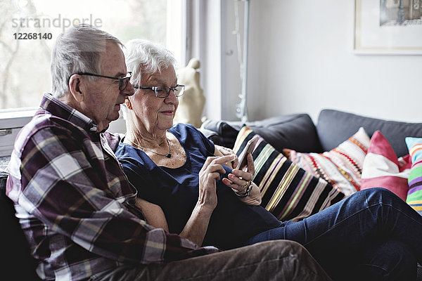 Senioren-Pärchen im Ruhestand teilen sich ein Smartphone auf dem Sofa im Wohnzimmer zu Hause.