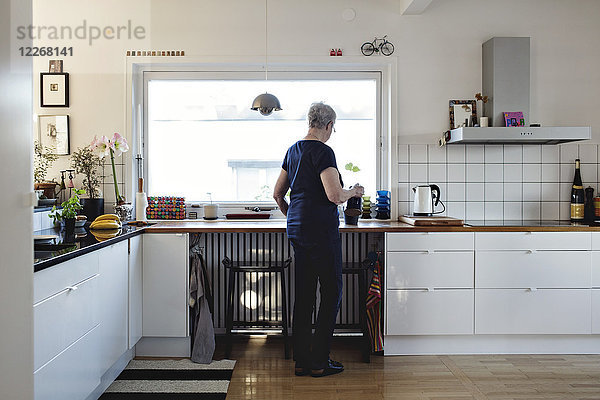 Durchgehende Rückansicht der älteren Frau  die in der Küche zu Hause steht.