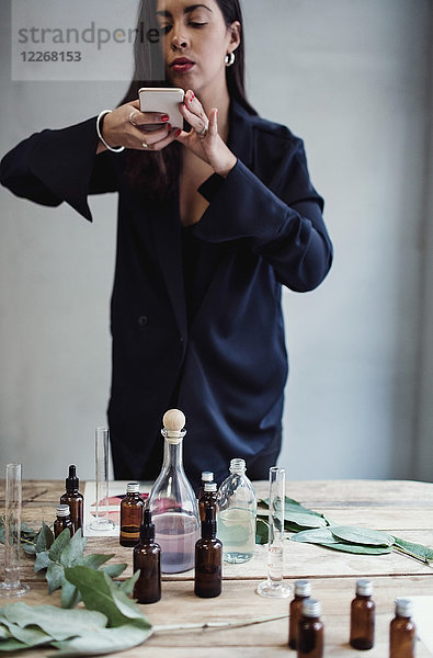 Mittlere erwachsene Unternehmerin beim Fotografieren von Parfümflaschen mit Smartphone auf dem Tisch gegen die Wand in der Werkstatt.