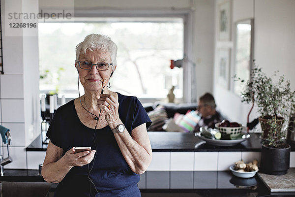 Zuverlässige Seniorin mit Smartphone beim Hören von In-Ear-Kopfhörern in der heimischen Küche