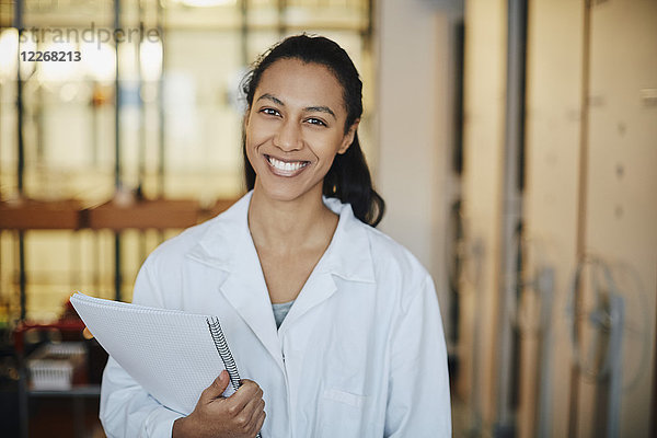 Porträt eines lächelnden jungen Chemiestudenten im Laborkittel stehend mit Buch in der Universität