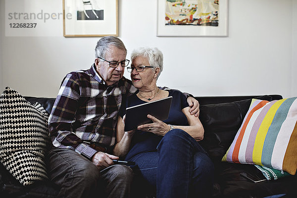 Seniorenpaar sitzt auf dem Sofa und teilt sich zu Hause ein digitales Tablett gegen die Wand.