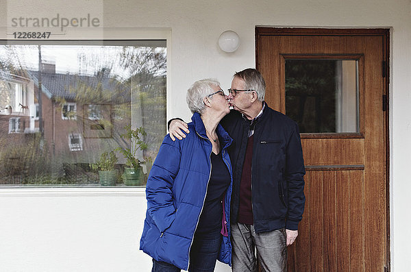 Seniorenpaar im Ruhestand küssend  während es sich gegen das Haus stellt.