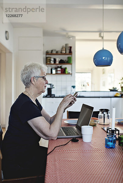 Seitenansicht der pensionierten Seniorin mit Smartphone im Sitzen mit Laptop am Esstisch