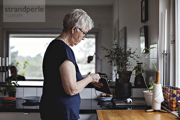 Seitenansicht der Seniorin beim Aufladen des digitalen Tabletts in der Küche zu Hause