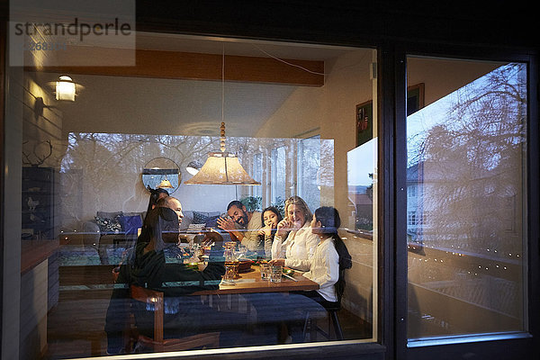 Glückliche Familie beim Abendessen am Tisch durch das Glasfenster bei Sonnenuntergang gesehen