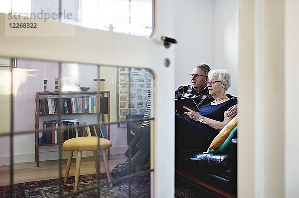 Volle Länge des Seniorenpaares auf dem Sofa mit digitalem Tablett von der Tür aus gesehen.