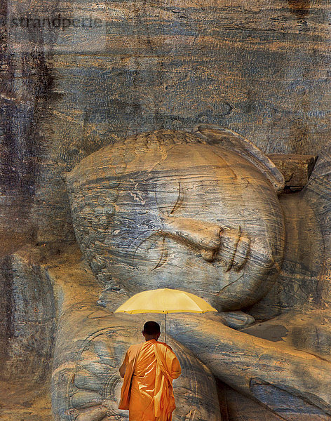 Rückansicht eines Mannes in orangefarbener Robe  der vor einer riesigen  liegenden  in den Fels gehauenen Buddha-Skulptur steht.