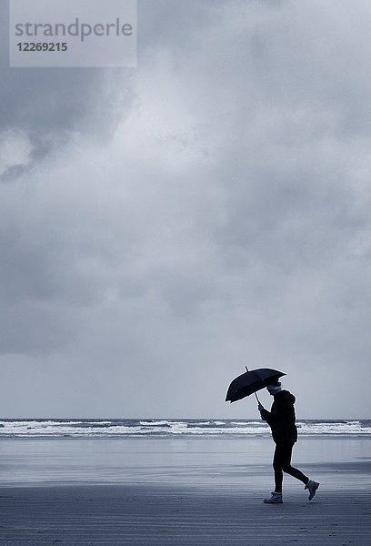 Seitenansicht eines Mannes mit Regenschirm  der unter stürmischem Himmel am Sandstrand am Meer entlang spaziert.