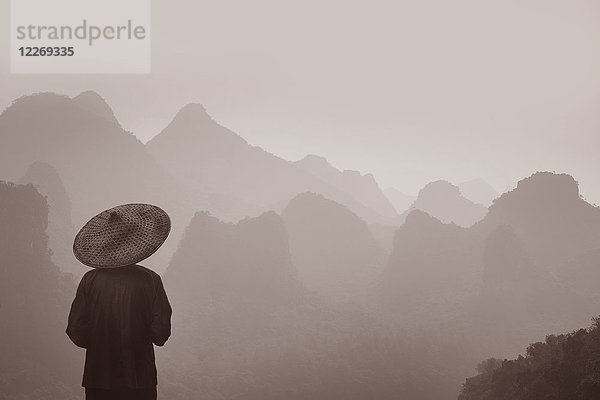 Rückansicht eines Mannes mit traditionellem Strohhut  der über die Bergkette blickt.