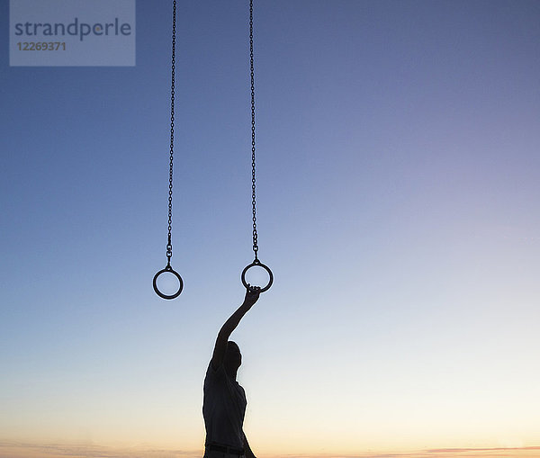 Silhouette eines Mannes  der bei Sonnenuntergang im Freien steht  den Arm hebt und sich an Gymnastikringen festhält.