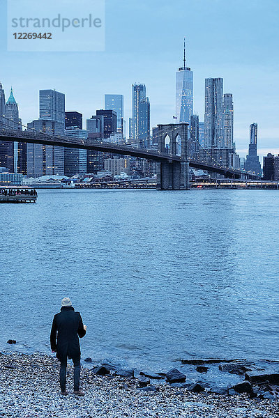 Mann blickt vom Flussufer auf die Brooklyn Bridge und die Skyline von Lower Manhattan  New York  USA