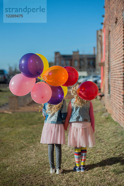 Zwei Schwestern halten sich mit einem Bündel bunter Luftballons an den Händen  Rückansicht