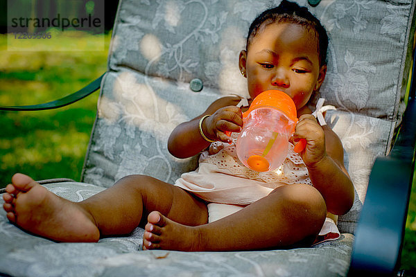 Babymädchen sitzt auf Gartensessel und trinkt aus Babybecher