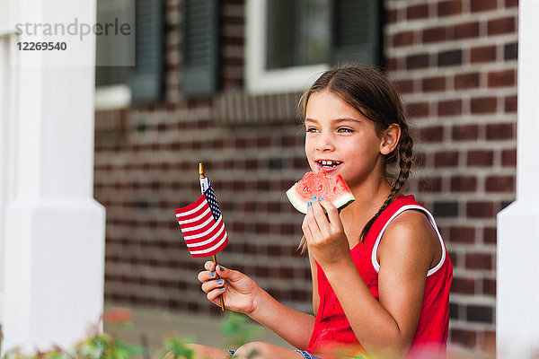 Junges Mädchen im Freien  hält kleine amerikanische Flagge und isst eine Scheibe Wassermelone