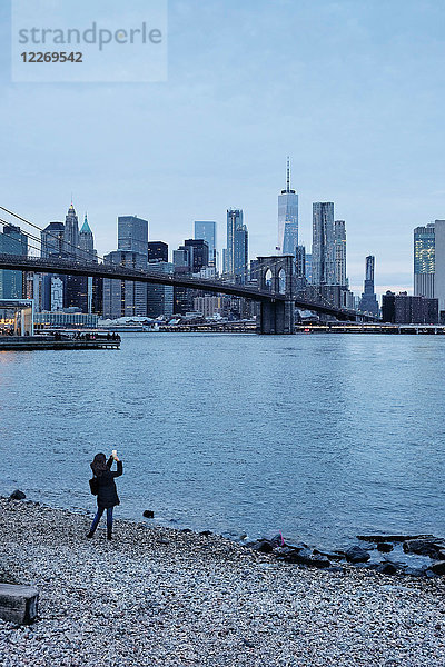 Frau fotografiert die Brooklyn Bridge und die Skyline von Lower Manhattan vom Flussufer aus  New York  USA