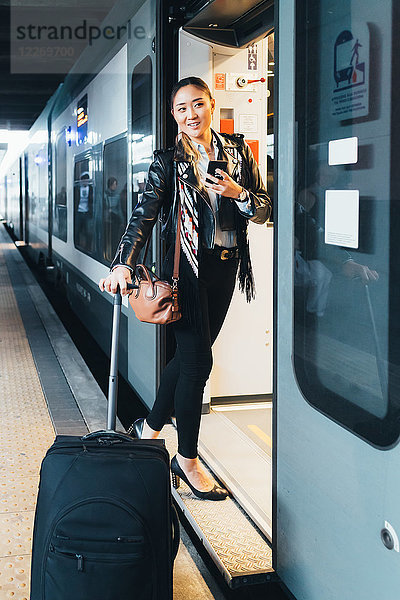 Frau steigt in den Zug ein  zieht einen Koffer mit Rädern  hält ein Smartphone