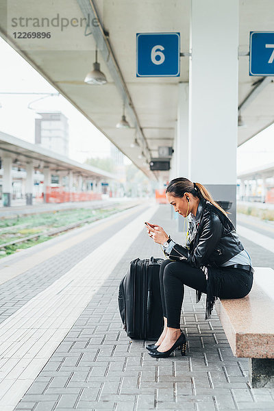 Frau sitzt auf Bahnsteig  benutzt Smartphone  Koffer neben ihr