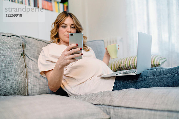 Schwangere Frau auf dem Sofa mit Laptop und Smartphone