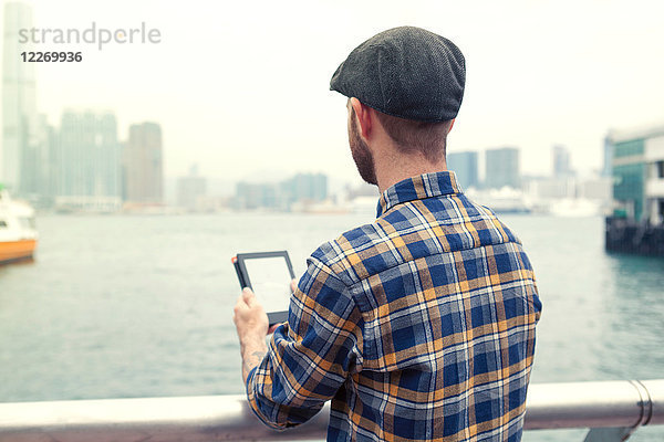 Mann hält digitales Tablett in der Hand und schaut weg  Blick auf Hafen  Rückansicht  Hongkong  China  Ostasien