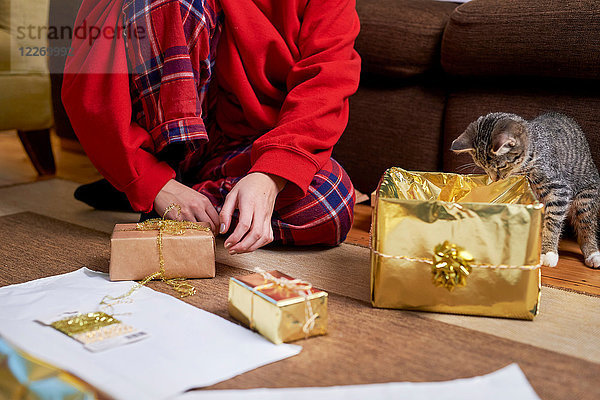 Junge Frau sitzt auf dem Wohnboden und packt mit neugieriger Katze Geschenke ein  Hals abwärts