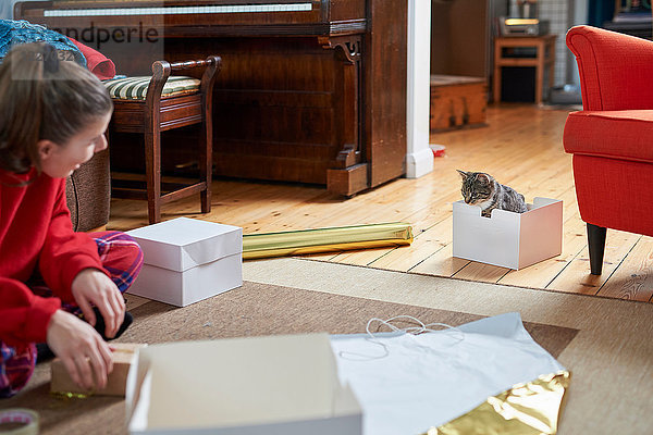 Junge Frau sitzt auf Wohnzimmerboden  packt Geschenke ein und beobachtet Katze