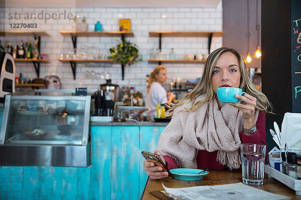 Frau sitzt im Café  hält ein Smartphone in der Hand und trinkt Kaffee