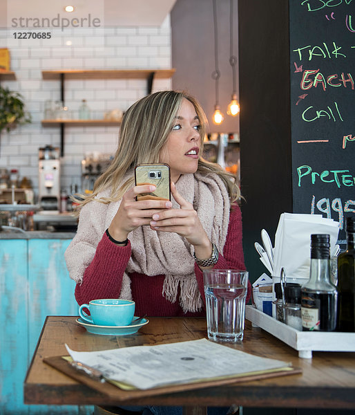 Frau sitzt im Café  hält ein Smartphone in der Hand und schaut weg