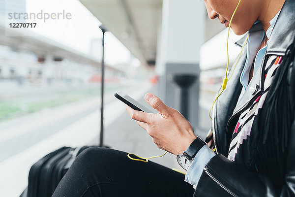 Frau sitzt auf dem Bahnsteig  benutzt ein Smartphone  trägt Kopfhörer  Mittelteil