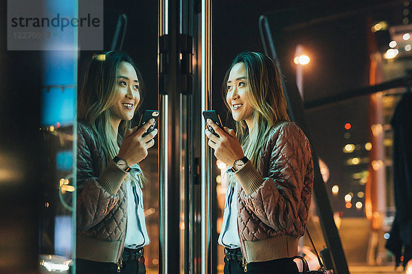 Frau steht im Freien  nachts  Smartphone in der Hand  Spiegelung im Fenster