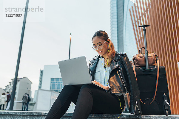 Geschäftsfrau  im Freien sitzend  Laptop benutzend  Rollkoffer neben ihr