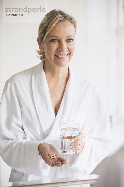 Lächelnde reife Frau im Bademantel  die am Badezimmerspiegel Vitamine einnimmt