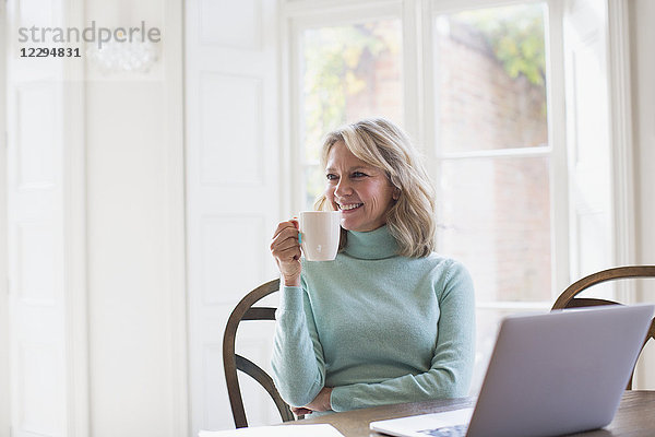 Lächelnde reife Freiberuflerin trinkt Tee und arbeitet am Laptop zu Hause