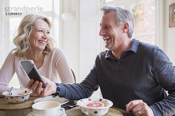 Lachendes reifes Paar  das ein Smartphone benutzt und frühstückt
