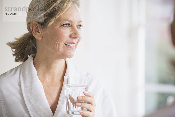 Lächelnde reife Frau im Bademantel trinkt Wasser