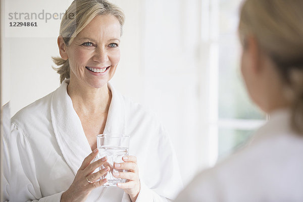 Lächelnde  selbstbewusste reife Frau im Bademantel  die am Badezimmerspiegel Wasser trinkt