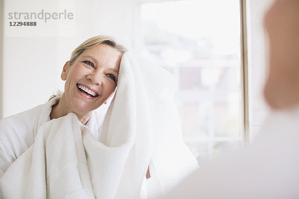 Lächelnde reife Frau trocknet ihr Gesicht mit einem Handtuch am Badezimmerspiegel