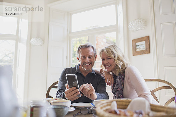 Lächelndes reifes Paar benutzt Smartphone am Esstisch