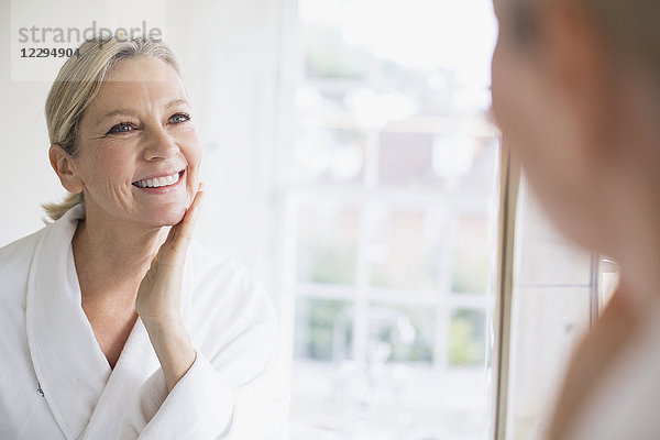 Lächelnde reife Frau berührt Gesicht im Badezimmerspiegel