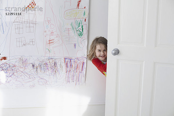 Porträt eines fröhlichen Mädchens  das sich hinter der Tür versteckt  gegen das Zeichnen an der Wand.