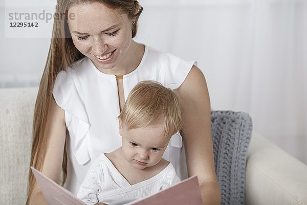 Lächelnde Mutter und Tochter beim Lesen des Bilderbuchs auf dem Sofa zu Hause
