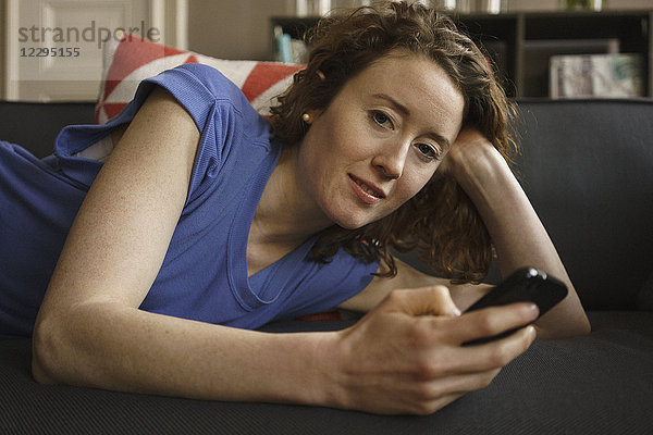 Porträt einer auf dem Sofa liegenden Frau mit dem Handy zu Hause