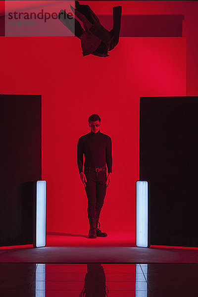 Volle Länge des jungen Mannes  der unter der herabfallenden Jacke inmitten der beleuchteten Lichter im roten Studio steht.