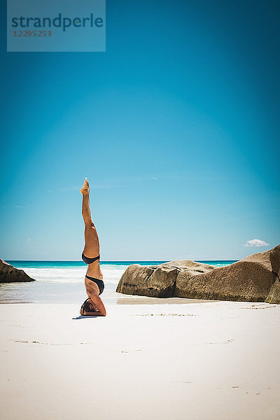 Mittlere erwachsene Frau im Bikini beim Kopfstand am Strand gegen den klaren blauen Himmel bei Sonnenschein