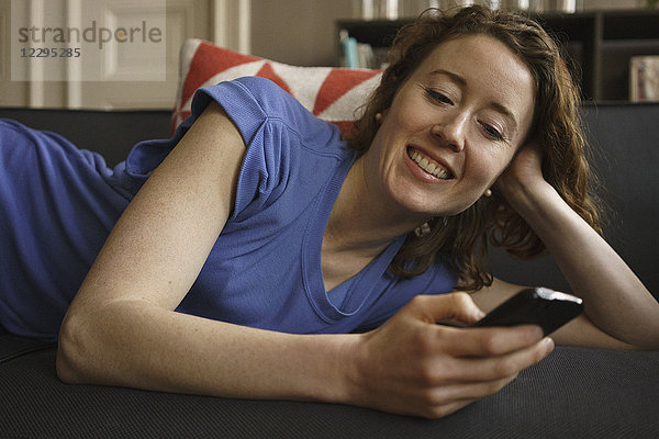 Lächelnde Frau auf dem Sofa liegend mit Smartphone zu Hause