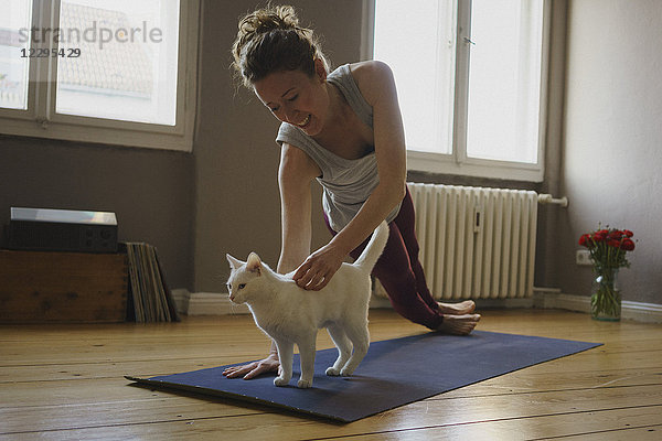Lächelnde Frau streichelt weiße Katze  während sie zu Hause Yoga übt.