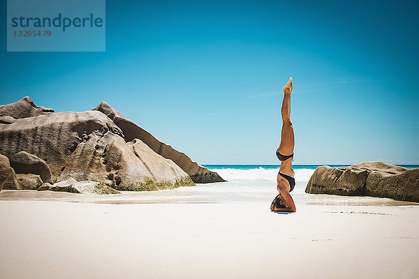 Seitenansicht der mittleren erwachsenen Frau im Bikini beim Kopfstand am Strand gegen den klaren blauen Himmel  Seychellen